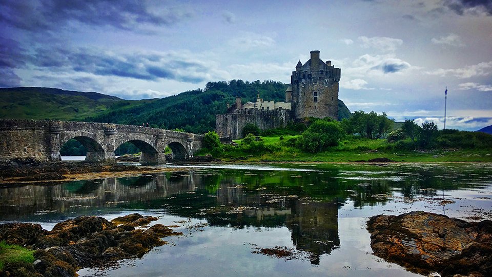 Le célèbre château d'Eilean Donan se reflétant dans l'eau, avec son pont de pierre, sous un ciel dramatique des Highlands.