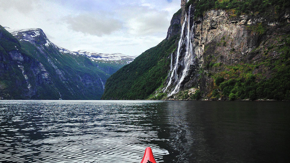 La pointe d'un kayak rouge naviguant sur les eaux sombres du Geirangerfjord, face aux majestueuses cascades des Sept Sœurs.
