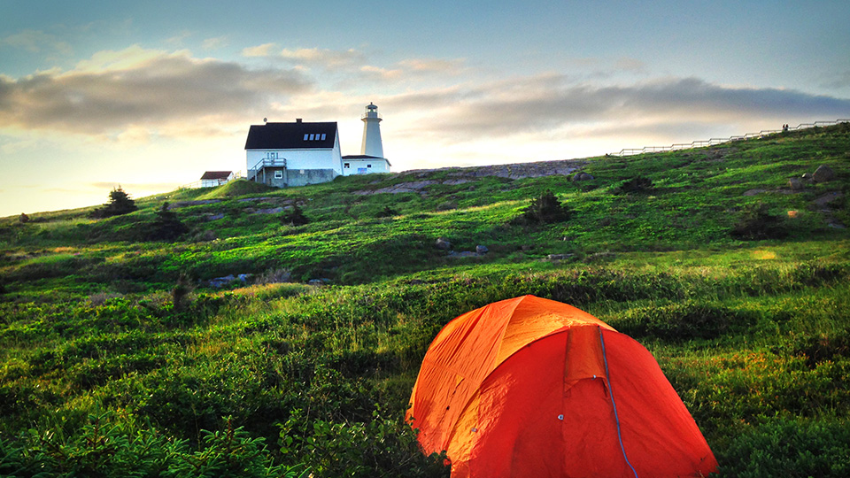 Une tente de camping orange plantée dans l'herbe verte au lever du soleil, avec le phare historique de Cape Spear en arrière-plan.